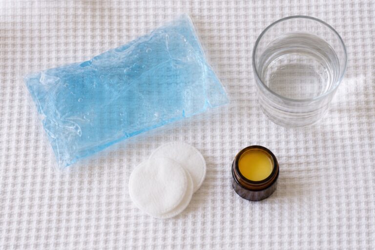 Flat lay of gel cold pack, water glass, cotton pads and arnica jar on a white towel