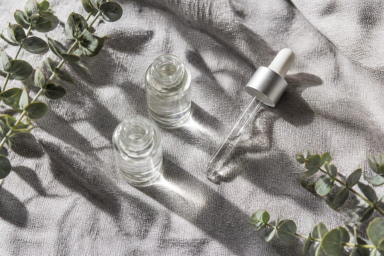 Flat lay of clear serum vials and glass dropper on grey linen with eucalyptus